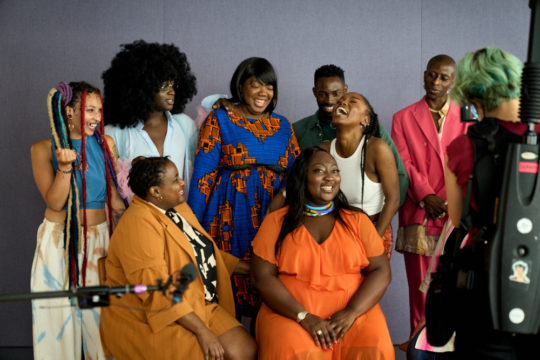 A group of people are casually posed in front of a backdrop. A boom mic and photographer can be seen in the foreground.